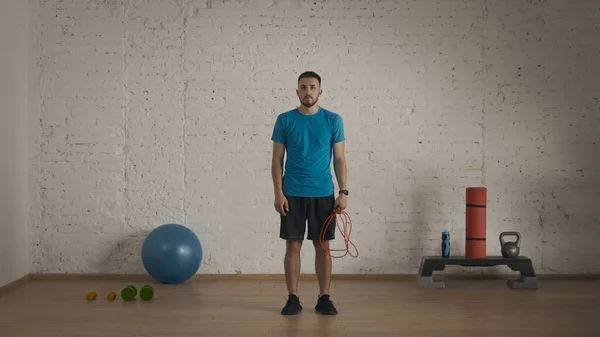 Sport indoors. Athletic fitness coach doing exercise at the home studio for online classes. Man in sportswear standing with skipping rope posing at the camera.
