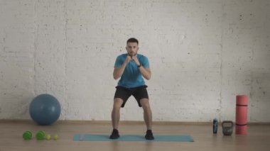 Evde kişisel spor eğitimleri. Atletik erkek fitness koçu çevrimiçi dersler için ev stüdyosunda egzersiz yapıyor. Spor kıyafetli bir adam çömelme hareketleri yapıyor ve tekniği açıklıyor. Sağlık sigortası konsepti. HDR