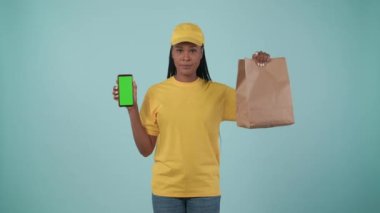 Kurye hizmeti konsepti. Sarı şapkalı ve tişörtlü bir teslimatçı kadının portresi. Elinde kese kağıdı ve akıllı telefon vardı. Paket servis ürünleri. Mavi arka planda izole edilmiş. Reklam alanı, çalışma alanı modeli.