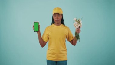 Kurye hizmeti konsepti. Sarı şapkalı ve tişörtlü bir teslimatçı kadının portresi. Elinde bir buket çiçek ve akıllı telefon vardı. Mavi arka planda izole edilmiş. Reklam alanı, çalışma alanı modeli.