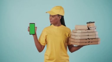 Kurye hizmeti konsepti. Sarı şapkalı ve tişörtlü bir teslimatçı kadının portresi. Elinde pizza kutuları, kahve ve akıllı telefon var. Mavi arka planda izole edilmiş. Reklam alanı, çalışma alanı modeli.