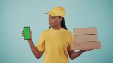 Kurye hizmeti konsepti. Sarı şapkalı ve tişörtlü bir teslimatçı kadının portresi. Elinde bir yığın posta kutusu ve akıllı telefon var. Mavi arka planda izole edilmiş. Reklam alanı, çalışma alanı modeli.