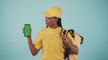 Kurye hizmeti konsepti. Sırt çantalı buzdolabı ve akıllı telefonlu sarı şapkalı teslimatçı kadının portresi onaylıyor. Mavi arka planda izole edilmiş. Reklam alanı, çalışma alanı modeli.