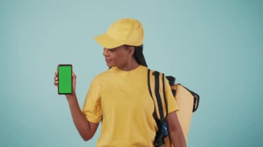Kurye hizmeti konsepti. Sırt çantalı bir kadın portresi buzdolabı ve akıllı telefonlu gülümseyen, baş parmağını gösteren. Mavi arka planda izole edilmiş. Reklam alanı, çalışma alanı modeli.