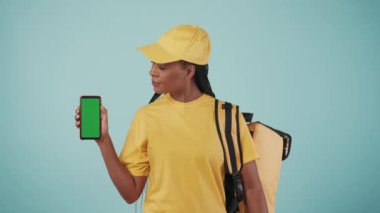 Kurye hizmeti konsepti. Sırt çantası buzdolabı ve akıllı telefonuyla kameraya gülümseyen, baş parmağını gösteren bir teslimatçı kadının portresi. Mavi arka planda izole edilmiş. Reklam alanı, çalışma alanı modeli.