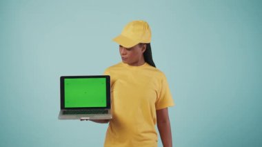 Kurye hizmeti konsepti. Sarı şapkalı ve tişörtlü bir kadının portresi dizüstü bilgisayar ve banka kartı taşıyor, kameraya gülümsüyor. Mavi arka planda izole edilmiş. Reklam alanı, çalışma alanı modeli.