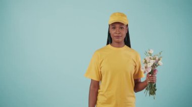 Kurye hizmeti konsepti. Sarı şapkalı ve tişörtlü bir kadının portresi elinde çiçek buketi, baş parmağını gösteriyor. Mavi arka planda izole edilmiş. Reklam için alan.