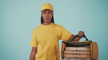 Kurye hizmeti konsepti. Sarı şapkalı ve tişörtlü bir kadının portresi kahve tutacağı ve pizza kutularıyla dolu buzdolabı çantası taşıyor. Mavi arkaplanda izole.