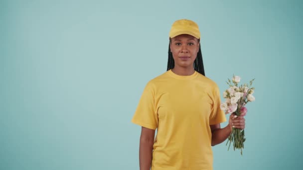 宅配サービスのコンセプト 黄色のキャップとTシャツの配達女性の肖像画は 花束を保持し 笑顔と親指を示しています 青い背景で孤立した 広告を挿入するスペース — ストック動画