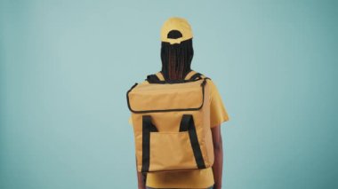 Kurye hizmeti konsepti. Sarı şapkalı, sırt çantalı, buzdolabı çantalı kadın portresi, kameraya dönüp başparmaklarını kaldırıyor ve gülümsüyor. Mavi arkaplanda izole.