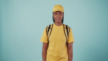 Kurye hizmeti konsepti. Sırt çantalı, çantalı, sarı şapkalı ve tişörtlü teslimatçı kadının portresi. Kameraya veriyor. Mavi arkaplanda izole.