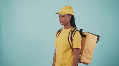Kurye hizmeti konsepti. Sırt çantalı, çantalı, sarı şapkalı ve tişörtlü teslimatçı kadının portresi onu müşteriye veriyor. Mavi arkaplanda izole.