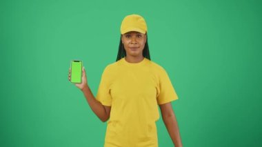 Kurye hizmeti konsepti. Sarı şapkalı ve tişörtlü bir teslimatçı kadının portresi. Elinde kese kağıdı ve akıllı telefon vardı. Yeşil arka planda izole edilmiş. Reklam alanı, çalışma alanı modeli.