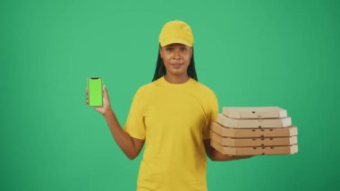 Kurye hizmeti konsepti. Sarı şapkalı ve tişörtlü pizza kutuları ve akıllı telefon taşıyan bir kadın portresi. Yeşil arka planda izole edilmiş. Reklam alanı, çalışma alanı modeli.