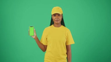 Kurye hizmeti konsepti. Banka kartı ve akıllı telefon taşıyan sarı şapkalı ve tişörtlü bir teslimatçı kadının portresi. Yeşil arka planda izole edilmiş. Reklam alanı, çalışma alanı modeli.