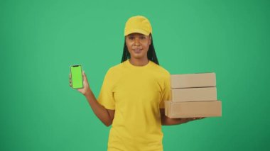 Kurye hizmeti konsepti. Sarı şapkalı ve tişörtlü bir teslimatçı kadının portresi. Elinde bir yığın posta kutusu ve akıllı telefon var. Yeşil arka planda izole edilmiş. Reklam alanı, çalışma alanı modeli.
