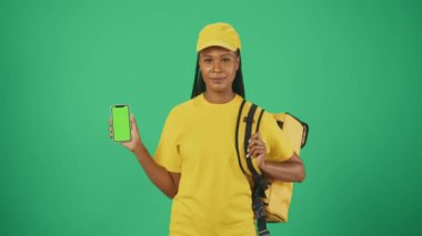Kurye hizmeti konsepti. Sırt çantalı buzdolabı ve akıllı telefonlu sarı şapkalı bir teslimatçının portresi. Yeşil arka planda izole edilmiş. Reklam alanı, çalışma alanı modeli.