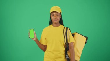 Sırt çantalı buzdolabı ve akıllı telefonlu sarı şapkalı bir teslimatçının portresi. İşaret ediyor, onay veriyor. Reklam alanı, çalışma alanı modeli. Yeşil arkaplanda izole.