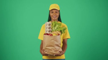 Kurye hizmeti konsepti. Sarı şapkalı ve tişörtlü teslimatçı kadının portresi. Elinde poşet dolusu yiyecek ve gülümseme var. Yeşil arkaplanda izole.