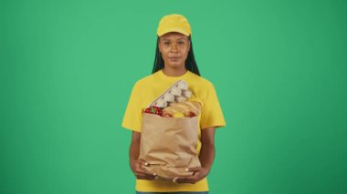 Kurye hizmeti konsepti. Sarı şapkalı ve tişörtlü teslimatçı kadının portresi. Bakkal ürünleriyle dolu bir kesekağıdıyla kameraya gülümsüyor. Yeşil arkaplanda izole.