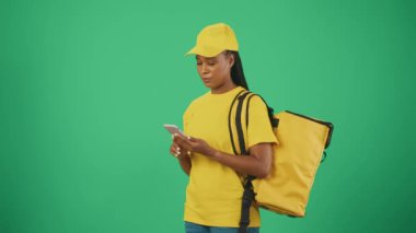 Kurye hizmeti konsepti. Sarı şapkalı ve tişörtlü akıllı telefon ve buzdolabı taşıyan teslimatçı kadının portresi müşteriye mesaj yazıyor. Yeşil arkaplanda izole.