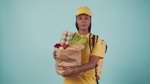 快递服务的概念 配送妇女的画像 头戴黄帽 头戴T恤 背着手提包 手里拿着装有杂货的纸袋 与蓝色背景隔离 — 图库视频影像