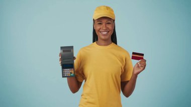 Portrait of a delivery woman in yellow cap and tshirt holding portable payment terminal and bank credit card, smiling. Isolated on blue background. clipart