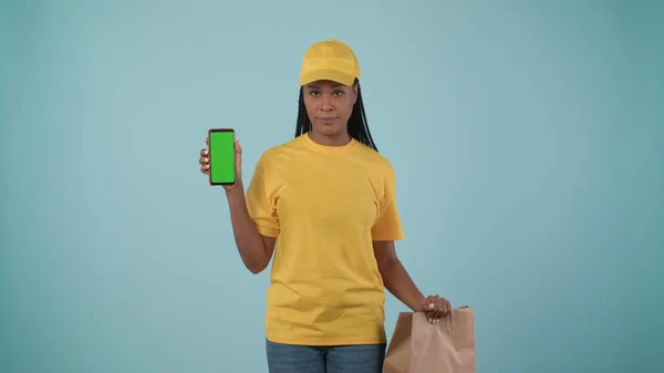 一个头戴黄帽 头戴T恤 手持纸袋和智能手机的送货妇女的画像 被蓝色背景隔离了广告区 工作空间模型 — 图库照片