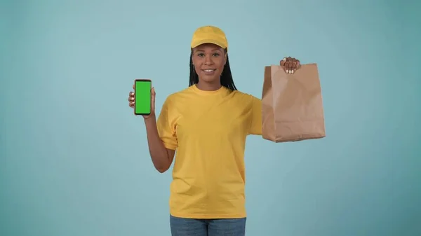 一个头戴黄帽 头戴T恤 手持纸袋和智能手机的送货妇女的画像 对着屏幕笑着 被蓝色背景隔离了广告区 工作空间模型 — 图库照片
