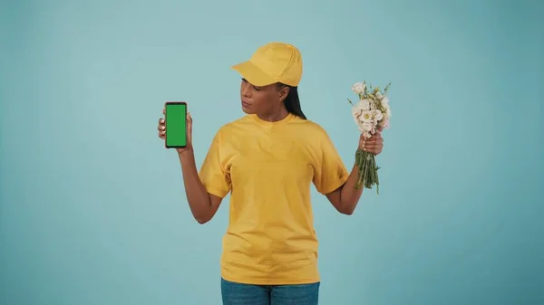 快递服务的概念 一个头戴黄帽 头戴T恤 手持一束鲜花和智能手机的送货妇女的画像 看着屏幕 广告区 工作空间模型 — 图库照片