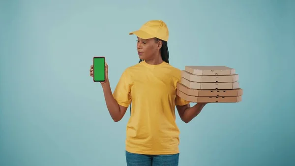 一个头戴黄帽 头戴T恤 手持披萨盒和智能手机的送货妇女的画像 看着屏幕 被蓝色背景隔离了广告区 工作空间模型 — 图库照片