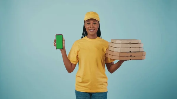 一个头戴黄帽 头戴T恤 拿着披萨盒和智能手机 对着相机微笑的送货妇女的画像 被蓝色背景隔离了广告区 工作空间模型 — 图库照片