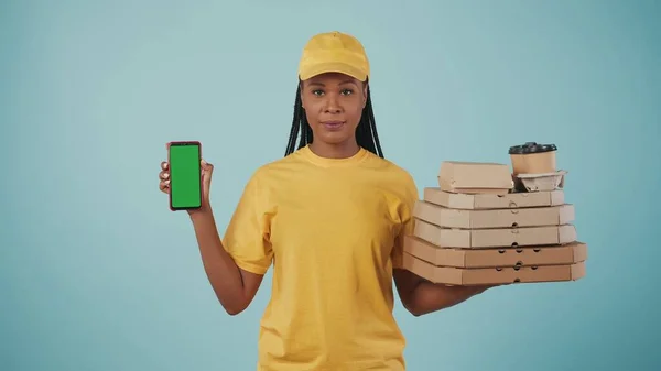 一个穿着黄色帽子和T恤的送货妇女拿着披萨盒 咖啡和智能手机 看着相机的画像 被蓝色背景隔离了广告区 工作空间模型 — 图库照片