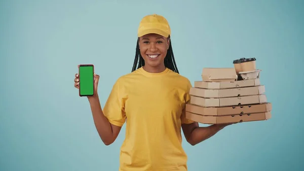 一个穿着黄色帽子和T恤的送货妇女拿着披萨盒 咖啡和智能手机 对着相机微笑的画像 被蓝色背景隔离了广告区 工作空间模型 — 图库照片