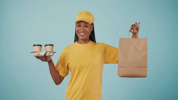 コーヒーカップホルダーと紙袋を持っている黄色い帽子とTシャツの配達女性の肖像画 青い背景に孤立した — ストック写真