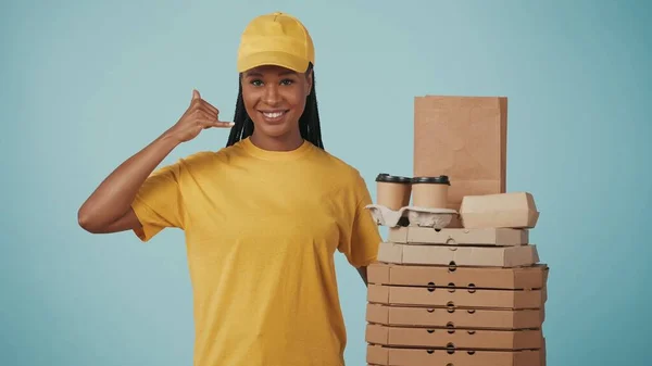 一个头戴黄帽 头戴T恤 提着食物背包的送货妇女的画像 在镜头前微笑 表现出呼叫手势 与蓝色背景隔离 — 图库照片