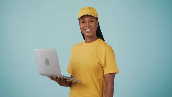 快递服务的概念 一个穿着黄色帽子和T恤 拿着笔记本电脑对着摄像机微笑的送货妇女的画像 与蓝色背景隔离 — 图库照片