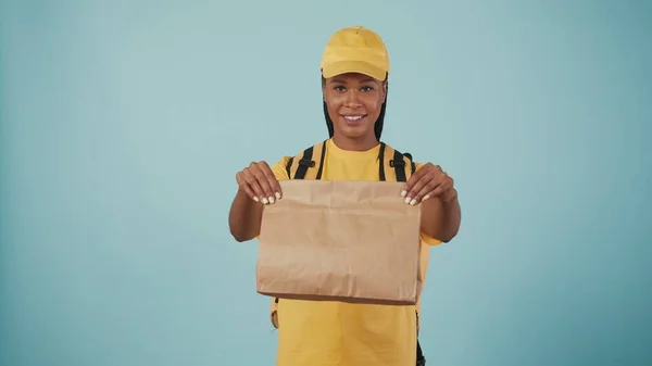 カメラに紙袋を与えるポータブルバックパック冷蔵庫付き黄色い帽子とTシャツの配達女性の肖像画 青い背景に孤立した — ストック写真