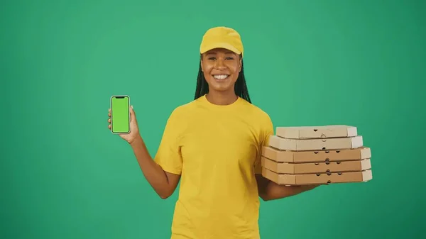 一个头戴黄帽 头戴T恤 拿着披萨盒和智能手机 对着相机微笑的送货妇女的画像 被绿色背景隔离 广告区 工作空间模型 — 图库照片