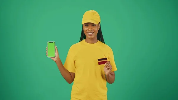 バンクカードとスマートフォンを持っている黄色いキャップとTシャツの配達女性の肖像画 緑色の背景に分離されています 広告エリア ワークスペースモックアップ — ストック写真