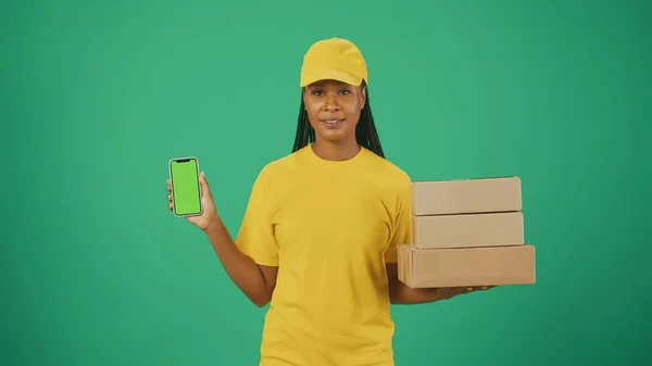 一个穿着黄色帽子和T恤 手持一堆堆盒子和智能手机的送货妇女的画像 被绿色背景隔离 广告区 工作空间模型 — 图库照片