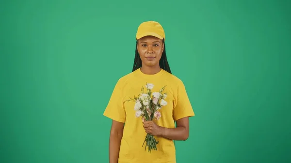 黄色い帽子とTシャツの配達女性の肖像画は カメラを見て花束を保持しています 緑の背景に孤立した — ストック写真
