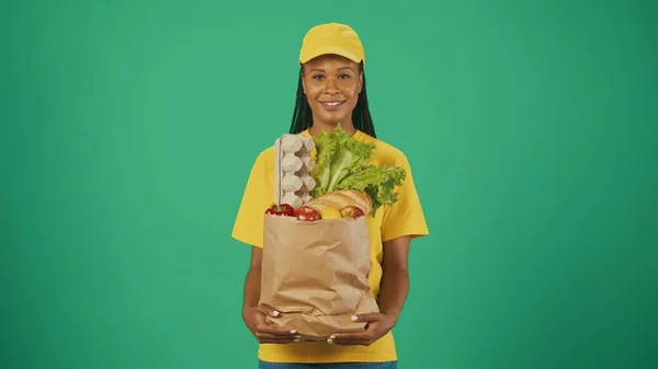 宅配サービスのコンセプト 紙袋食料品を保有する黄色い帽子とTシャツの配達女性の肖像画 緑の背景に孤立した — ストック写真