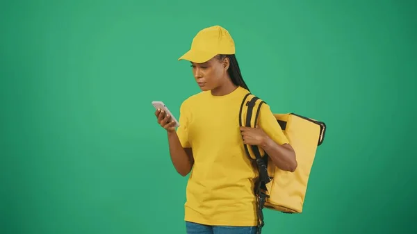 带有智能手机和背负式冰箱的黄色帽子和T恤的送货妇女的肖像 为客户写信息 被绿色背景隔离 — 图库照片