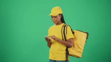 Kurye hizmeti konsepti. Sarı şapkalı ve tişörtlü akıllı telefon ve taşınabilir sırt çantalı kadın portresi. Mesaj kutusunu kontrol edip bekliyor. Yeşil arkaplanda izole.