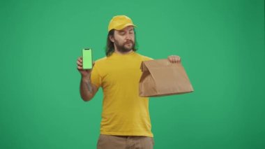 Kurye hizmeti konsepti. Sarı şapkalı ve tişörtlü bir teslimatçının portresi. Elinde kese kağıdı ve akıllı telefon vardı. Paket servis ürünleri. Yeşil arka planda izole edilmiş. Reklam alanı, çalışma alanı modeli.