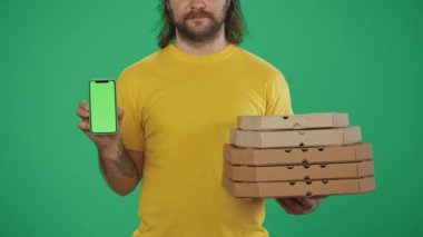 Yakın plan çekim. Sarı şapkalı bir teslimatçının portresi. Elinde bir yığın pizza kutusu ve akıllı telefon var. Paket servis ürünleri. Yeşil arka planda izole edilmiş. Reklam alanı, çalışma alanı modeli.