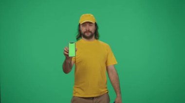 Kurye hizmeti konsepti. Elinde su şişesi ve akıllı telefon tutan sarı şapkalı bir teslimatçının portresi. Ürün teslimatı. Yeşil arka planda izole edilmiş. Reklam alanı, çalışma alanı modeli.