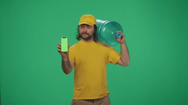 Kurye hizmeti konsepti. Sarı şapkalı bir teslimatçının portresi. Elinde büyük bir şişe ve akıllı telefon var. Ürün teslimatı. Yeşil arka planda izole edilmiş. Reklam alanı, çalışma alanı modeli.
