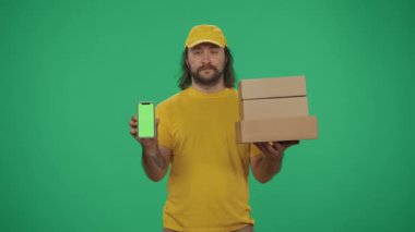 Kurye hizmeti konsepti. Sarı şapkalı bir teslimatçının portresi. Elinde bir yığın posta kutusu ve akıllı telefon var. Yeşil arka planda izole edilmiş. Reklam alanı, çalışma alanı modeli.